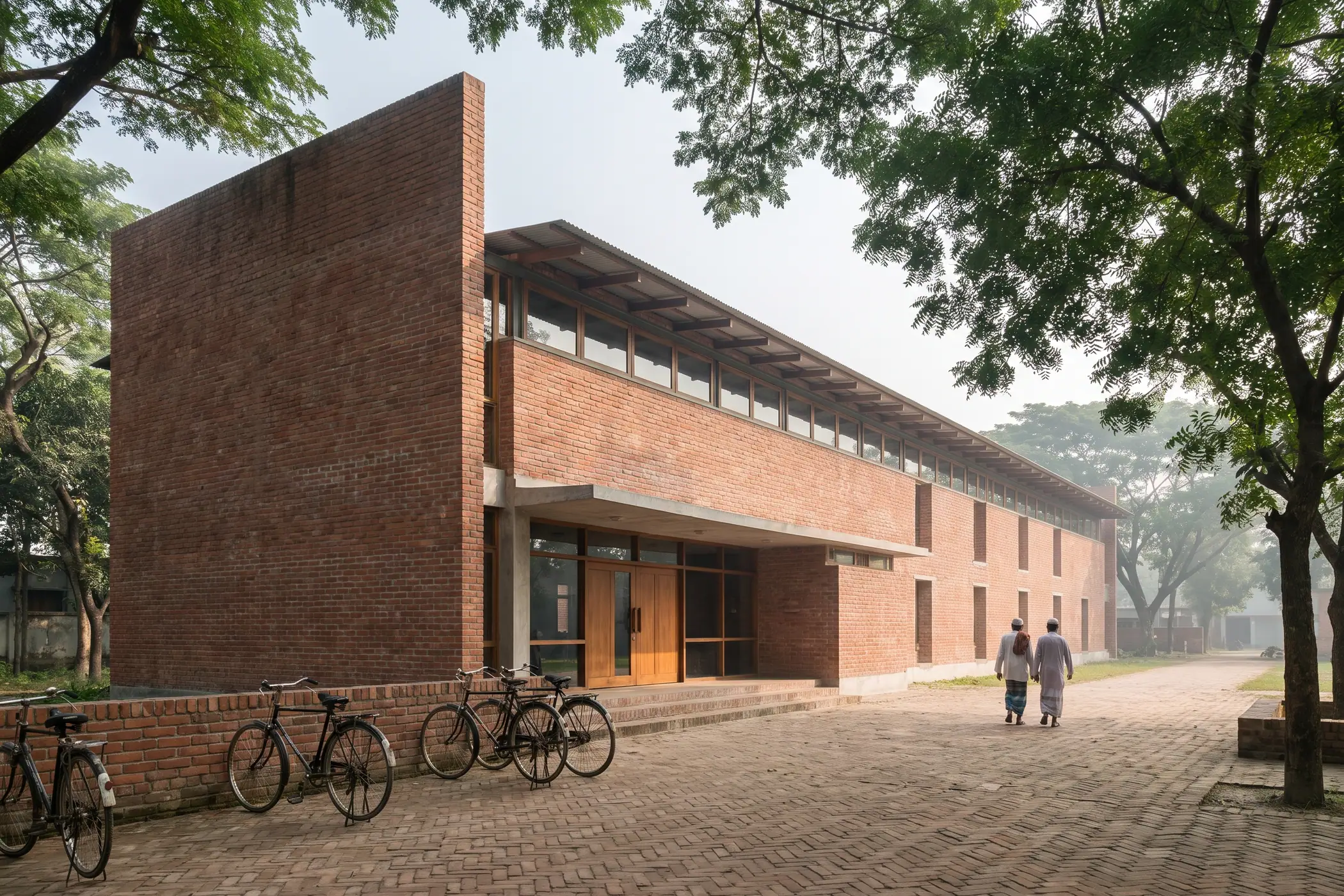 Community Library, Rajshahi: long two-storey red brick building with a clerestory under a timber-rafter roof, seen across a brick-paved forecourt shaded by rain trees
