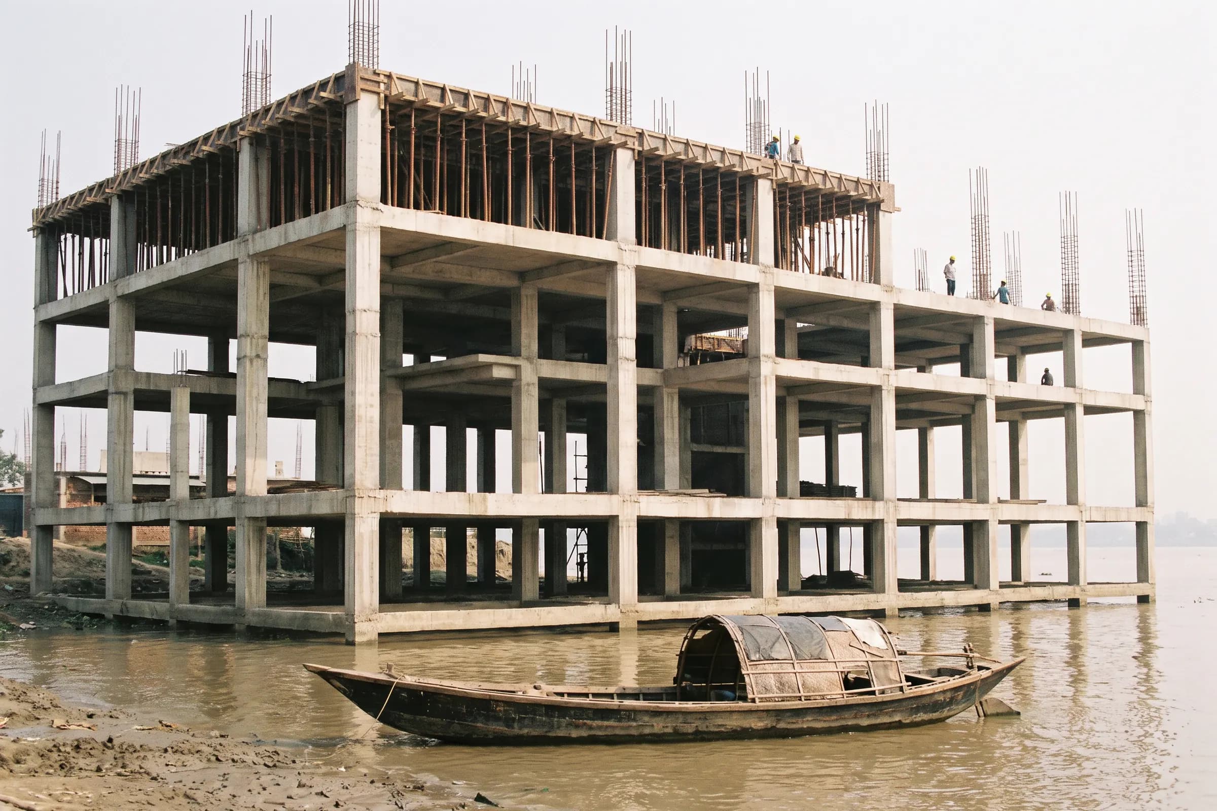 Concrete frame under construction on the riverbank, with a wooden boat in the foreground