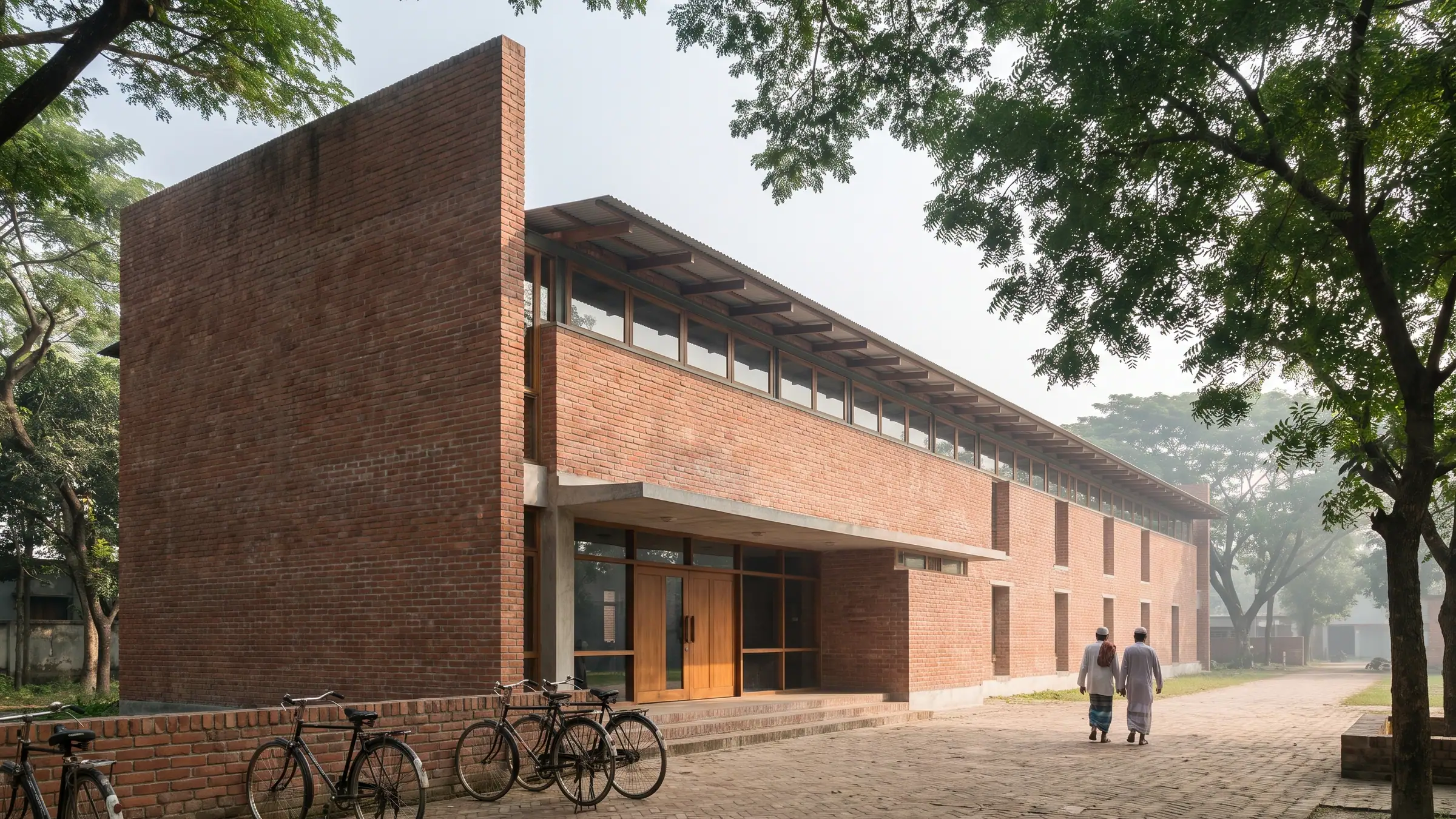 Community Library, Rajshahi: long two-storey red brick building with a clerestory under a timber-rafter roof, seen across a brick-paved forecourt shaded by rain trees